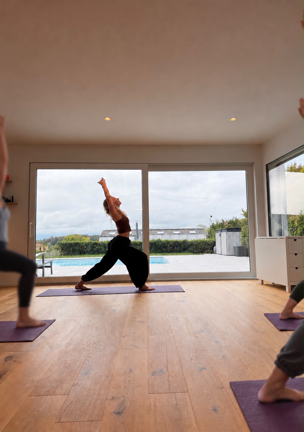 Participante en fente, bras levés vers le ciel, sur un tapis violet dans une salle lumineuse ouverte sur le jardin et la piscine