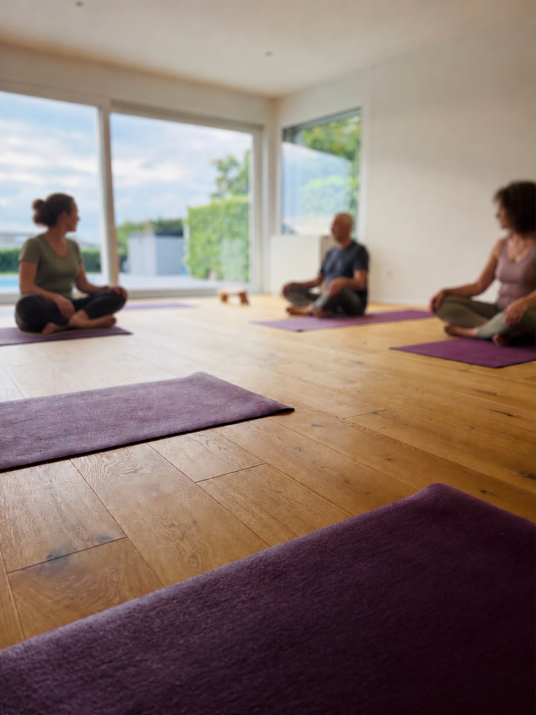 Trois personnes assises en tailleur sur des tapis violets dans une salle lumineuse ouverte sur le jardin